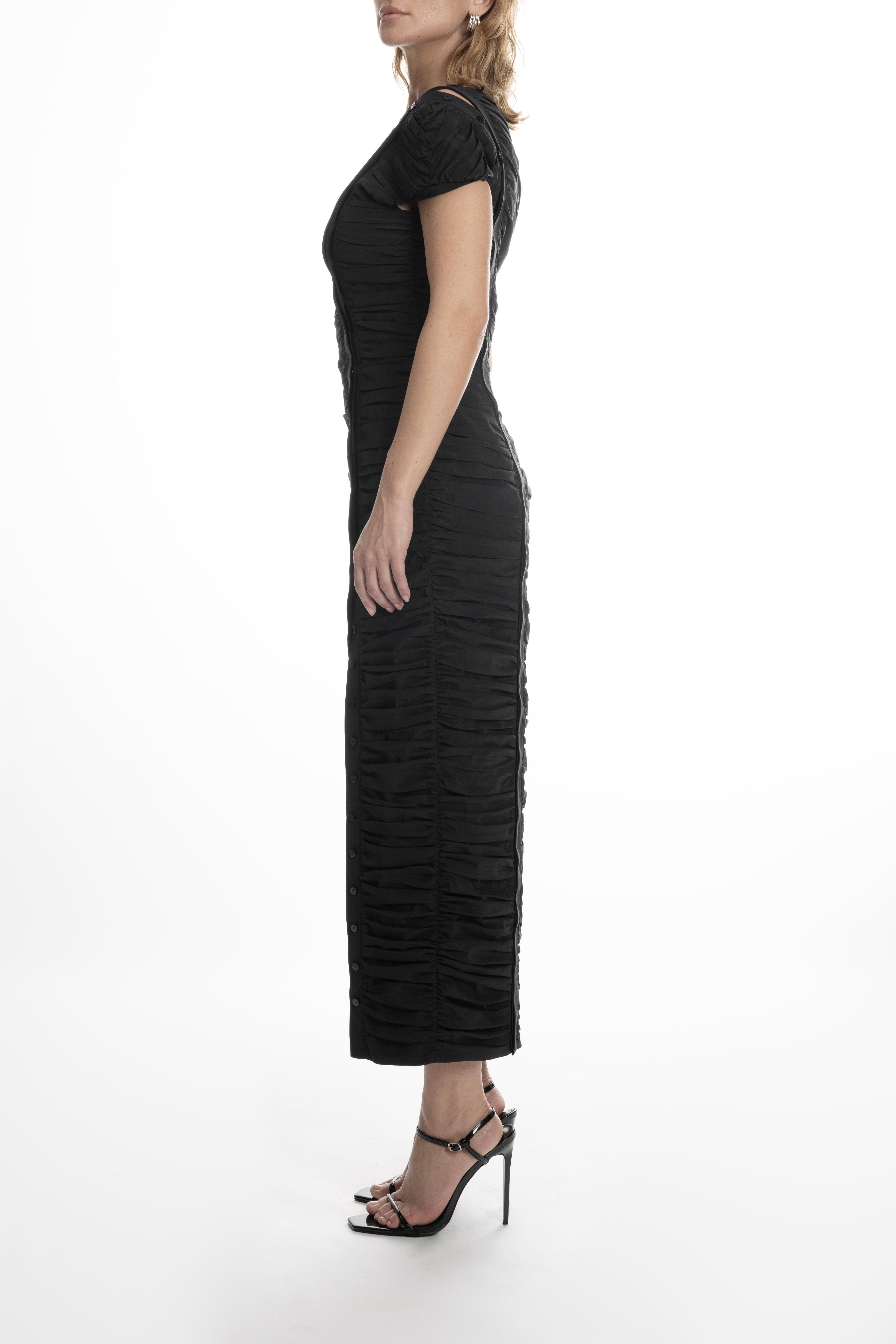 Side view of the black Pleated Dress showing sculptural pleating, fitted silhouette and mid-calf length