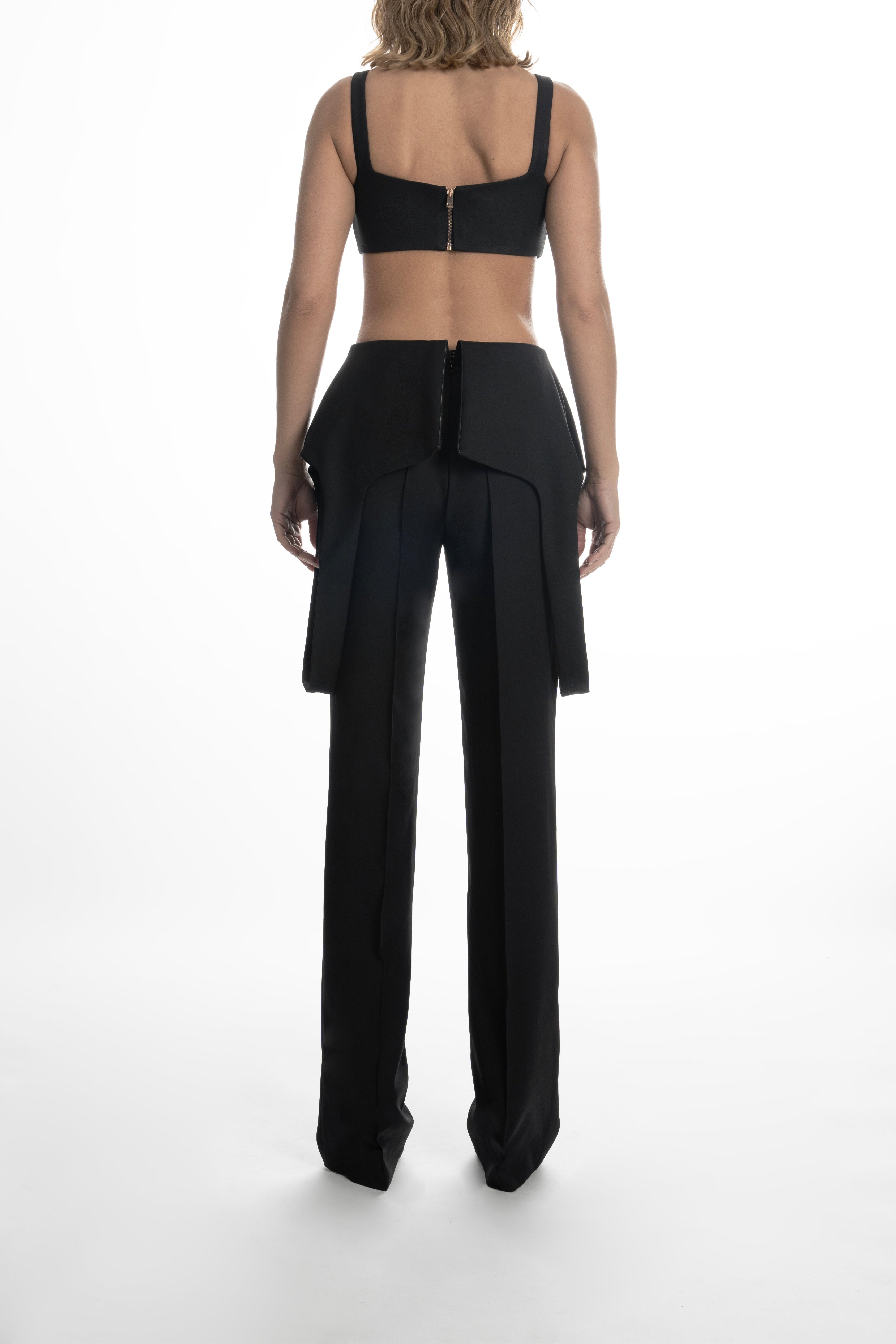 Woman full-length wearing black high-waist wool trousers with draped hip panels and straight-leg cut