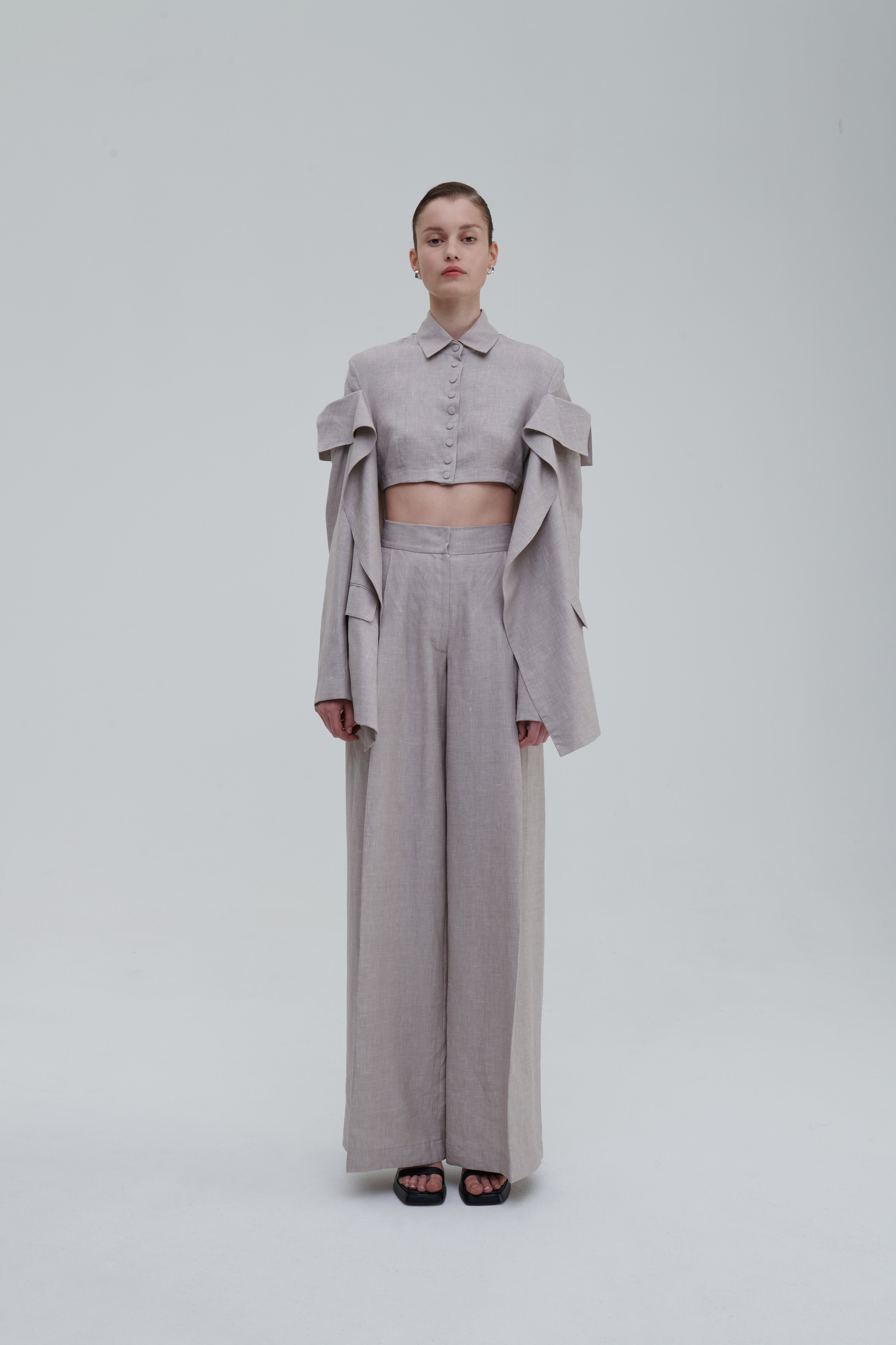 CLASSIC LINO TROUSERS by Sagio from the Spring/Summer 2025 collection, featuring wide-leg high-waisted linen pants in light beige, styled with a matching cropped top and statement sleeves. Discover more at thesagio.com.