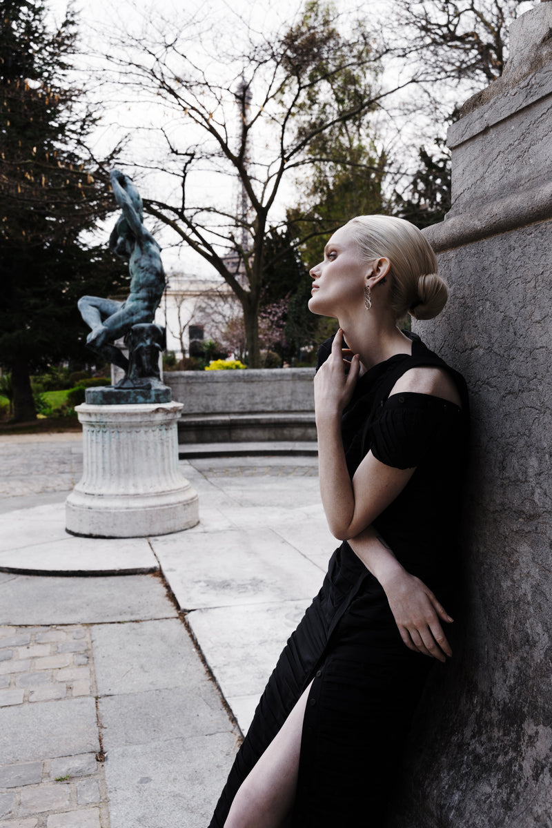 Model wearing the black 100%-silk Pleated Dress with sculptural gathers and a thigh-high front slit, leaning against stone in an outdoor setting
