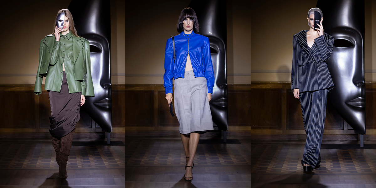 Three fashion models walk in a runway show wearing distinct outfits: an olive green cropped jacket with a brown draped skirt, a bright blue leather jacket with a light gray skirt, and a dark navy pinstripe blazer and matching trousers, set against a dark hall with large sculptural faces.