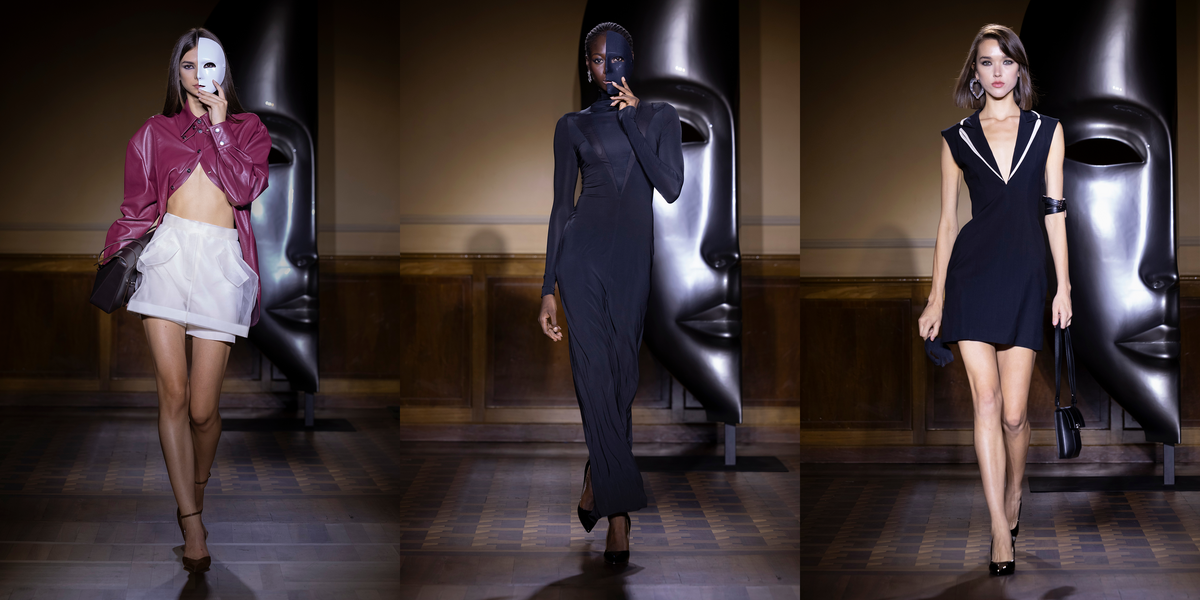 Three fashion models walk a runway in front of large black sculptural masks. The left model wears a white mask, a magenta leather jacket, a white mini skirt, and black heels.