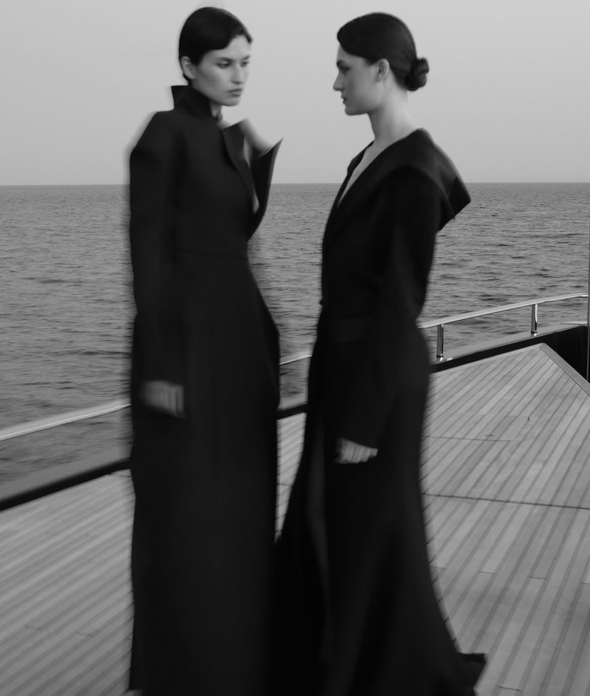 Two models in long black coats on a deck at sea
