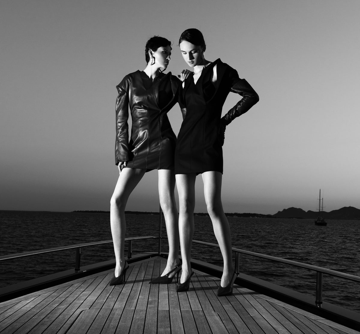 Black and white fashion photograph of two women standing on a wooden boat deck overlooking the sea at dusk, wearing short dark dresses.